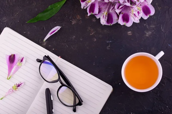 흰 컵, 아름다운 꽃, 검은 배경, 펜과 안경이 있는 노트북에 향기로운 차. — 스톡 사진