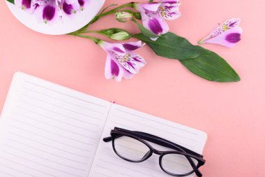 Beyaz bardak kokulu çay, güzel çiçekler, pembe bir arka plan, bir kalem ve gözlük ile bir defter.