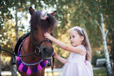 Pembe elbiseli küçük bir kız parkta kahverengi bir midilliyle..