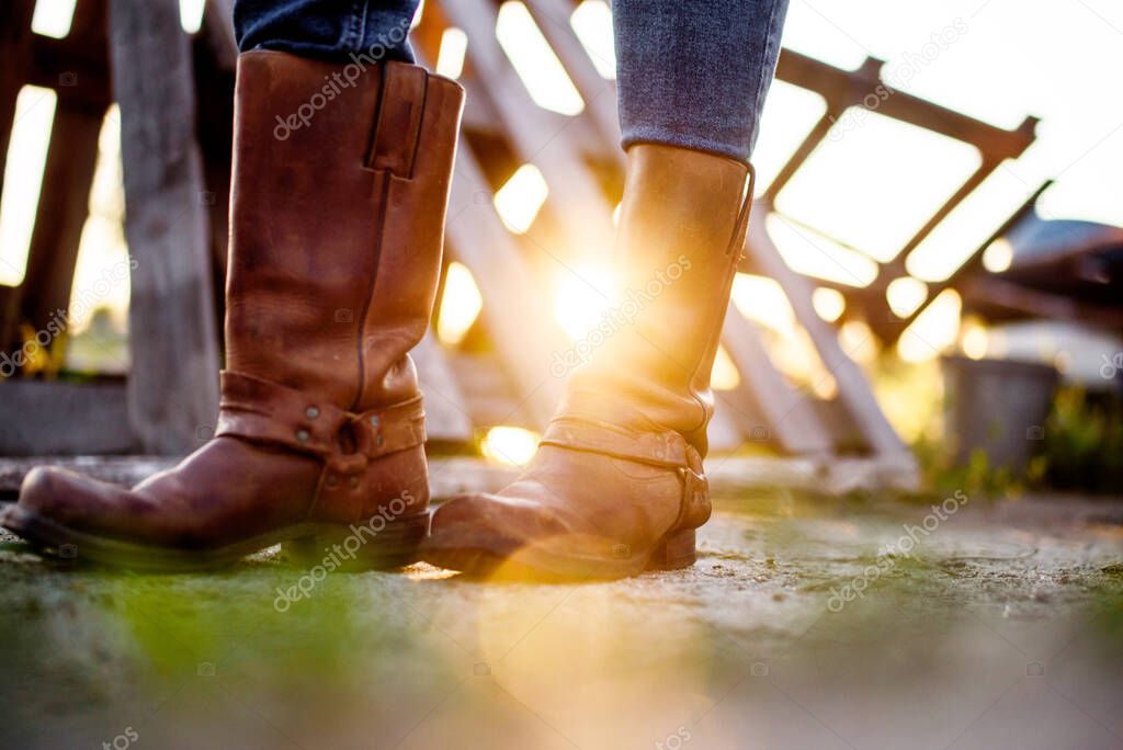 The girl stands on the ranch in cowboy boots and catches the rays of the setting sun. View from below.