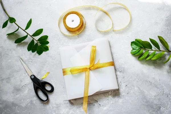 Συσκευασία ένα δώρο, σε όμορφο λευκό χαρτί, μια χρυσή κορδέλα. Σπαρίδες από πράσινα φυτά, ψαλίδια. Χειροποίητη συσκευασία — Φωτογραφία Αρχείου