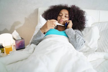 Grip evde yatak odasında hasta Afrikalı-Amerikalı kız. Onun ağzına bir termometre tutan soğuk arama Doktor telefonla, hasta genç siyah kadınla.