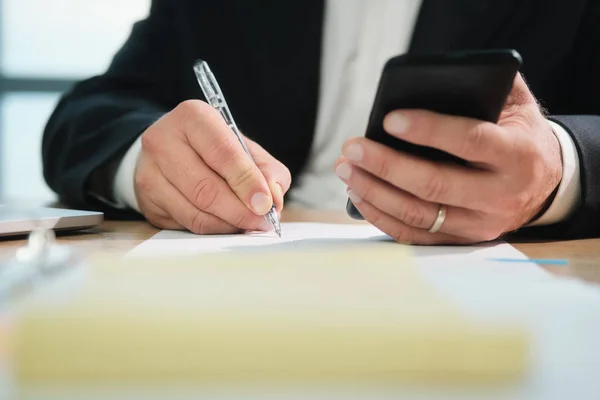 Manos del hombre de negocios escribiendo con la pluma y sosteniendo el teléfono —  Fotos de Stock