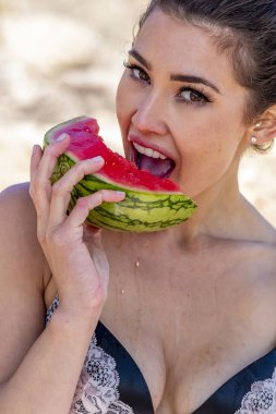 Güneşli bir günde kumsalda karpuz yiyen muhteşem bir esmer bikini mankeni.