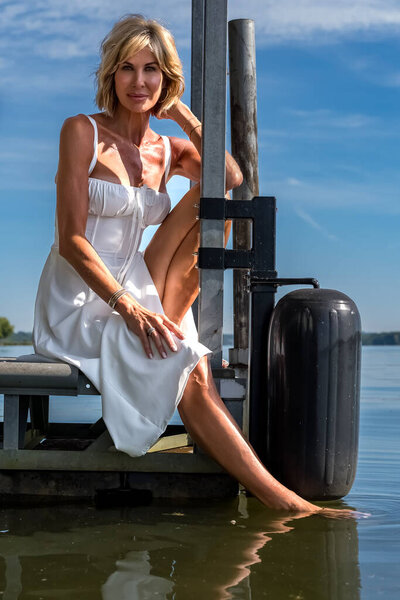 A beautiful blonde woman stands confidently, her radiant smile and poised demeanor exuding elegance. Her graceful stance highlights her charm, creating an image of timeless beauty and self-assurance.