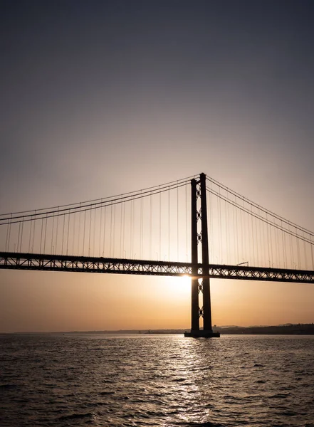 25 de Abril Bridge and the River Tagus at sunset. The silhouette of a backlit 25 April Bridge crossing the calm waters of the River Tagus. The landmark suspension bridge crosses the Portuguese river from Lisboa to Almada.