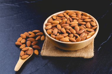Tahta kaşık ya da kepçeyle siyah taş masadaki bademler. Tahta kasede badem. Koyu tahtaya serilmiş fındıklar.