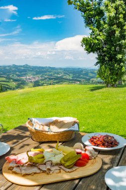Brettljause with cold cuts, beans salad and bread on wooden table with beautiful view over green meadow to panoramic south styrian wine route clipart