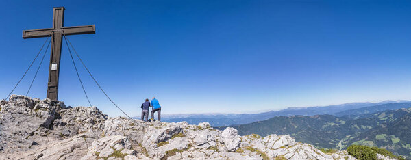 Два туриста стоят у креста и наслаждаются прекрасным видом с вершины горы Хохланч в Штирии, Австрия
