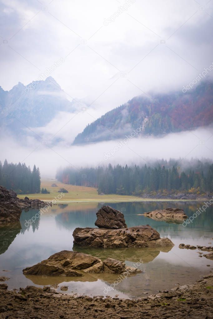 View over lake Laghi di Fusine on a foggy morning with some rocks at the beach and mountain range Mangart near Tarvisio in Italy