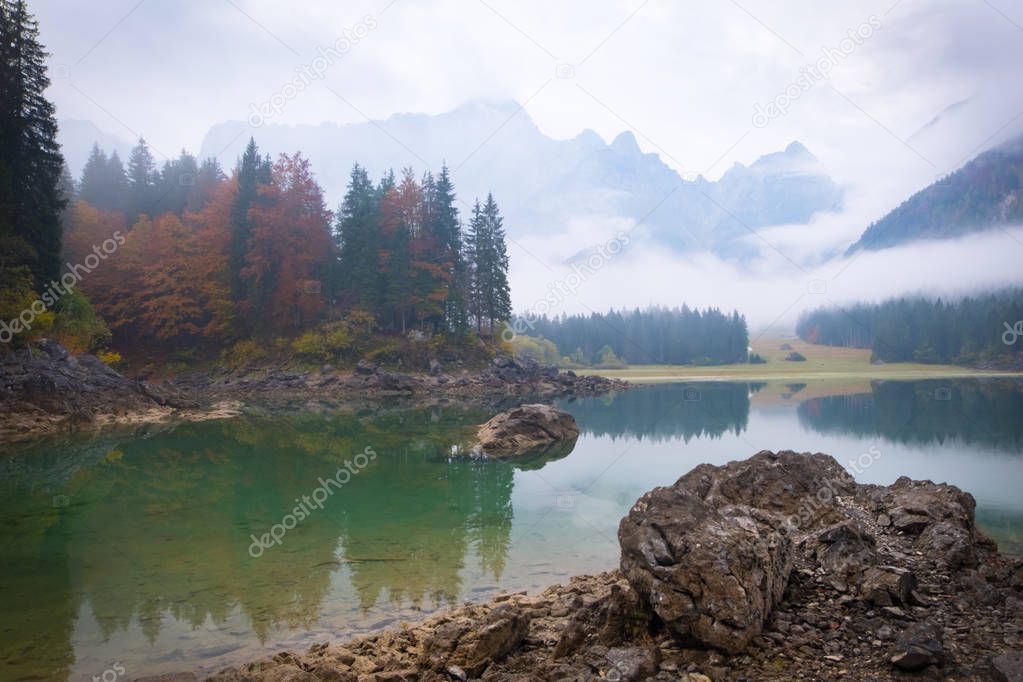 View over lake Laghi di Fusine on a foggy morning with some rocks at the beach and mountain range Mangart near Tarvisio in Italy
