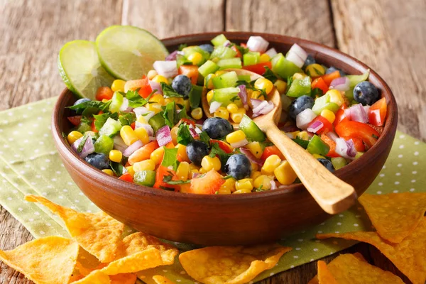 Mexican spicy salsa of corn, blueberries, pepper, herbs and onions close-up in a bowl and nachos chips on a table. horizonta