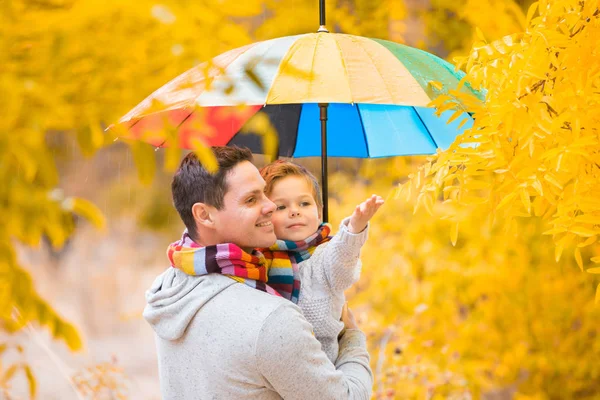小さな男の子と彼の父親はカラフルな傘の下にあります 暖かい服やカラフルなスカーフで家族 子供は手のひらで雨滴をキャッチします — ストック写真