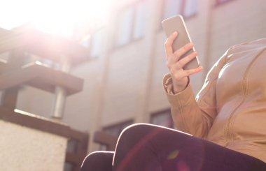 Gün ışığında cep telefonu kullanan rahat kız. Akıllı telefon dokunmak ve soğutma trendy kıyafetler şık genç kadınla. Güzel gün batımı veya gündoğumu şehir merkezinde. Modern kentsel yaşam tarzı.