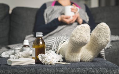 Cold medicine and sick woman drinking hot beverage to get well from flu, fever and virus. Dirty paper towels and tissues on table. Ill person wearing warm woolen stocking socks in winter. clipart