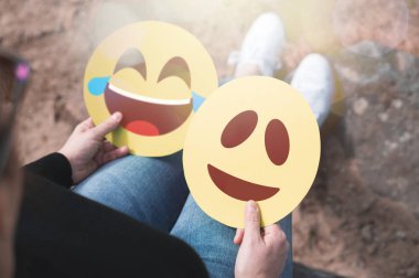 2 karton ifadeler elinde tutan kadın. Mutlu bir şekilde gülerek gülümseyen yüzler. İki modern iletişim ve ifade simgeleri kağıda basılmıştır. Mutluluk, sevinç ve duygu kavramı ifade.