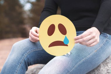 Sad paper emoticon in hand. Depressed woman holding printed crying smiley face and sitting on a rock. Modern communication and smiley icon on cardboard. Sadness, depression and emotions concept. clipart