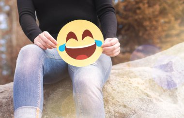 Laughing emoticon with tears of joy. Woman having fun outside and holding a cheerful printed paper smiley face. Happy communication and smiley icon on cardboard. Happiness, joy and expression concept. clipart