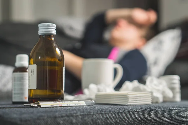 Sick woman holding head and forehead with hand and arm. Medicine, thermometer, hot beverage and dirty paper towels in front. Person having flu, fever and headache on sofa couch. Caught cold in winter.