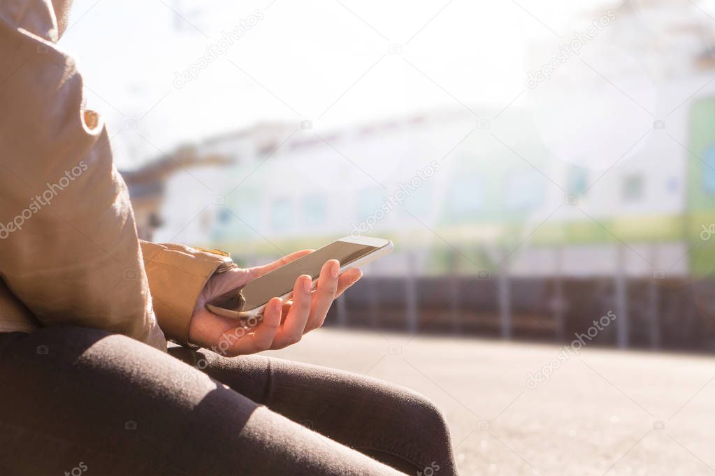 Woman waiting for train in platform and using smartphone to buy e-ticket or browsing schedule. Sunny day in the station with bright lens flare from the sky. Modern public transportation concept.