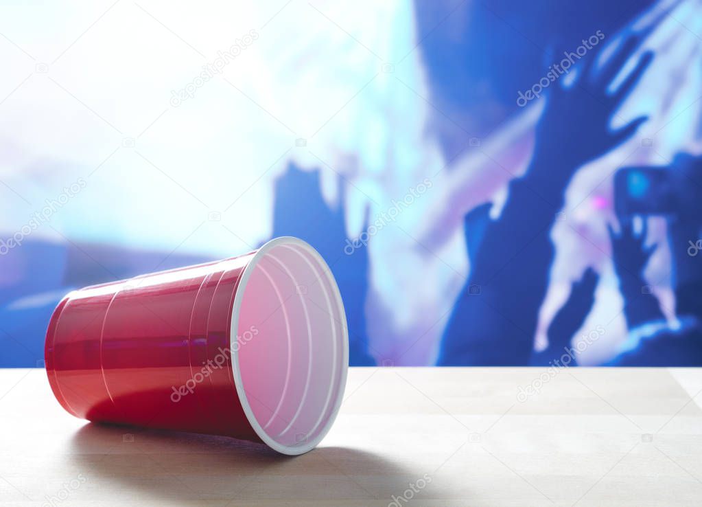Fallen plastic red party cup on its side on a table. Nightclub or disco full of people dancing on the dance floor in the background. Perfect for marketing and promotion for events or college fest.