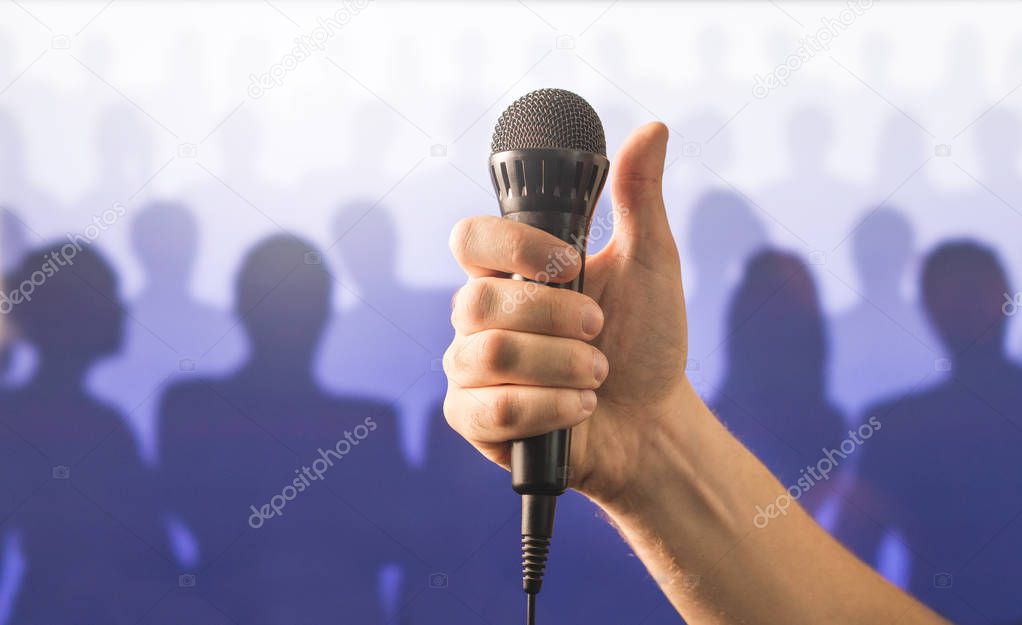 Hand holding microphone and showing thumbs up in front of a crowd of silhouette people. Public speaking and giving speech concept. Singing well live to mic, good karaoke or successful talent show.
