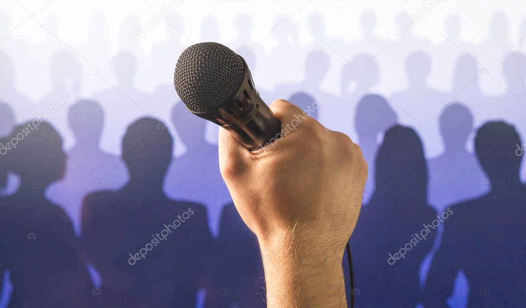 Public speaking and giving speech concept. Hand holding microphone in front of a silhouette audience and crowd of people. Singing to mic in karaoke or talent show concept.