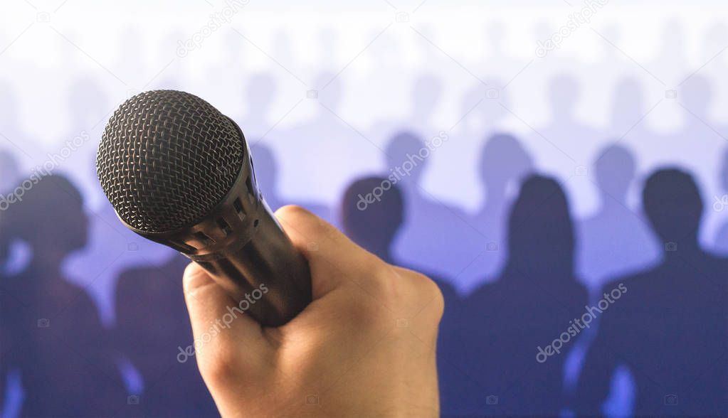 Public speaking and giving speech concept. Close up of hand holding microphone in front of a crowd of silhouette people. Singing to mic in karaoke or talent show concept.