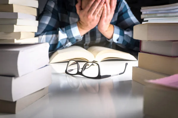 Stressed, tired and unhappy student. Too much work from school. Young man covering face with hands surrounded by stacks of books. Glasses on table.