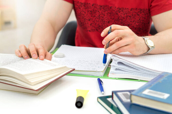 Young athletic man and student studying and writing notes in public or school library in college or university. Stack and pile of books, pen and paper on table. Negative copy space.