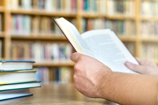 Man or student reading a book in public or school library in college or university. Education, studying and literature service concept. Hands holding book with bookshelf and bookcase in the background
