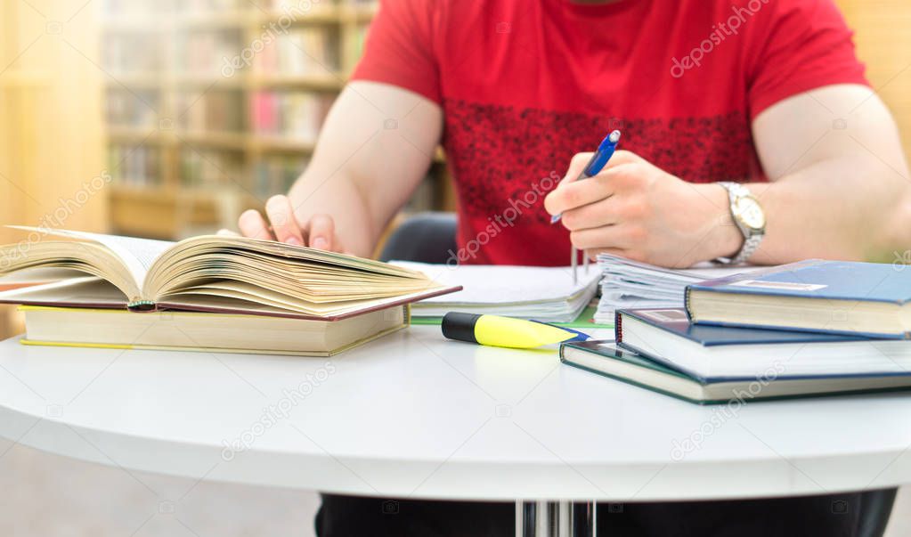 Young athletic man and student studying and writing notes in public or school library in college or university. Stack and pile of books, pen and paper on table. Negative copy space.