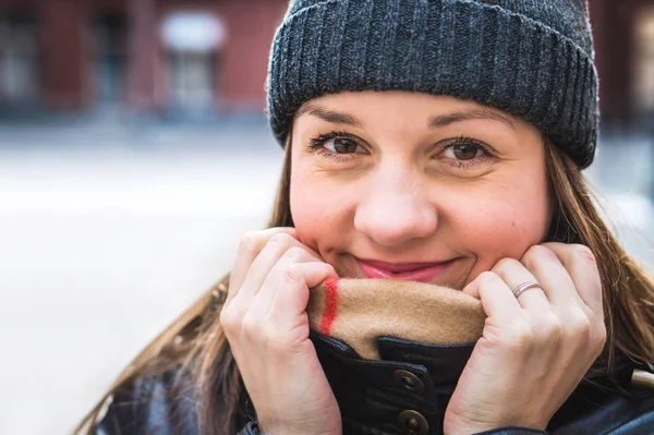 Cute woman wearing a beanie in winter. Happy and smiling person hiding behind scarf and holding collar with hands. Outdoor lifestyle portrait in city. Ring in finger.