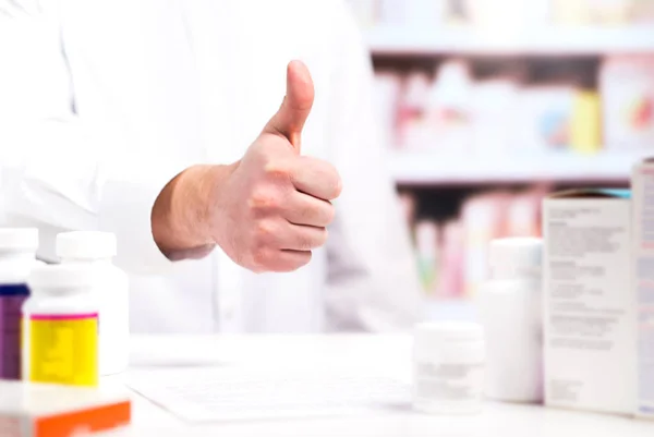 Happy pharmacist showing thumbs up at pharmacy counter full of medicine. Drugstore shelves in the background. Positive and cheerful druggist. Good or great customer service in medical store.