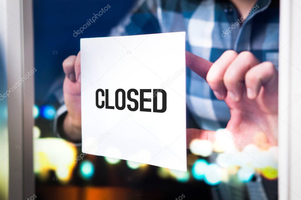 Bankruptcy, failed business going down or opening times concept. Man putting closed sign in window in cafe, restaurant, shop, store or agency. Late at night in city.