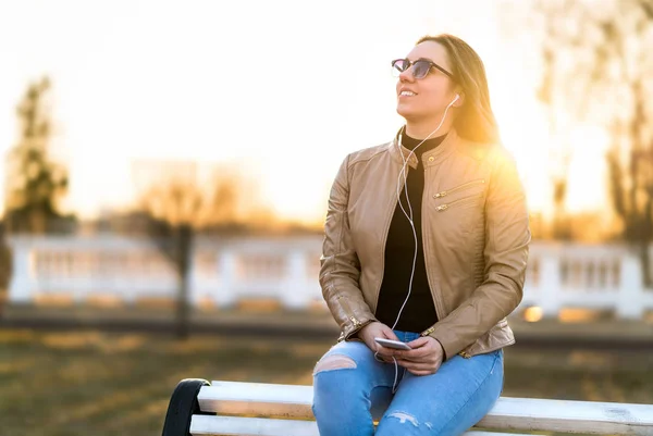 Woman listening to music outdoors. Happy smiling lady sitting on park bench holding mobile phone in sunset. Listening to audiobook outside.