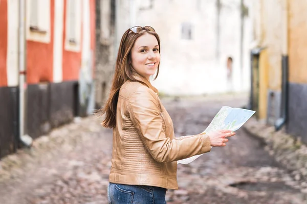 Woman with map looking straight to camera. Happy female traveler in city street or old town posing. Portrait of smiling person on vacation.