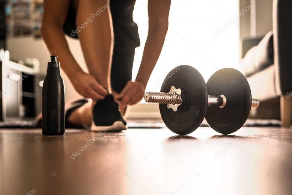 Morning workout routine in home gym. Fitness motivation and muscle training concept. Man in sneakers tying shoelaces in sunlight. Athlete starting exercise with dubbell weight.