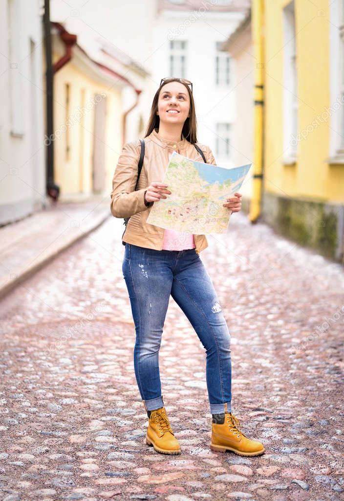 Vertical shot of young happy woman  holding map. Smiling traveler sightseeing in city. Tourist with backpack looking directions. Travel and navigation concept. Student on vacation.