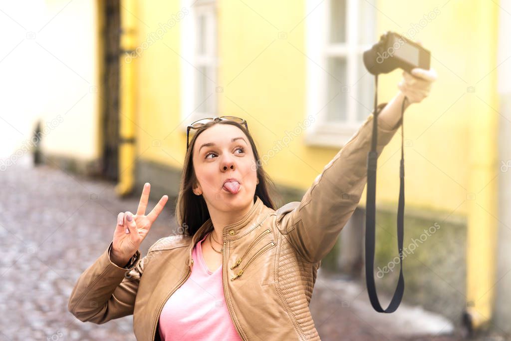 Young woman making silly face and showing tongue to camera while taking selfie. Happy person having fun and making v sign with hand. Girl taking funny photo.