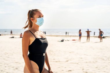 Corona virüsü sırasında sahildeki insanlar. Koronavirüsten korumak için maske takan bir kadın. Covid, seyahat, tatil ve turizm konsepti. Enfeksiyondan korunan bir turist. Yaz tatili.