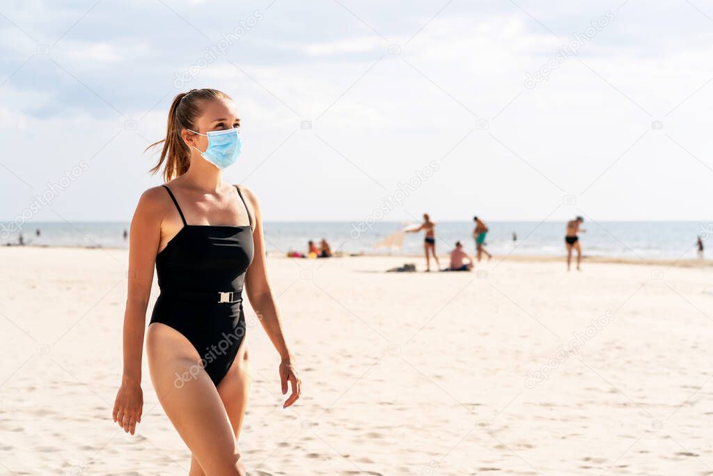 Woman walking on the beach wearing a face mask to protect from corona virus. Tourist on a vacation with people in the background. Facemask on mouth in public place. Safety during coronavirus, covid.