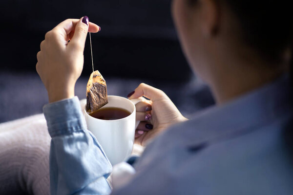 Woman holding tea bag in cup to make a healthy hot beverage in winter. Something warm for the cold season. Organic herbal teabag. Peaceful mood on weekend morning. Sick person with fever or flu.