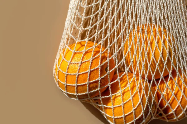 Ripe oranges in a white string bag. Beige background. Close up. Copy space.