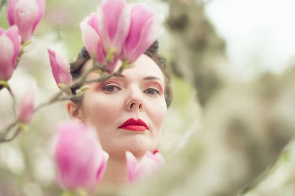 Portrait of a brunette with a red lipstick lips in a red dress near a blooming magnolia