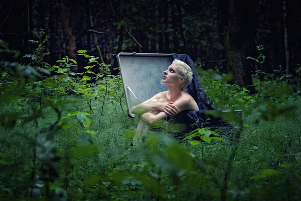 A young girl with short white hair is sitting in a large suitcase in the middle of the forest