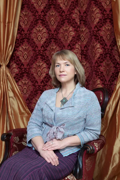 Портрет жінки в блакитній блузці та фіолетовій спідниці, що сидить у кріслі на бордовому фоні — стокове фото