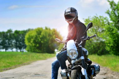 Motosikletçi kask ve deri ceketli motosikletine açık havada oturur. Motosiklet arka plan seyahat. 