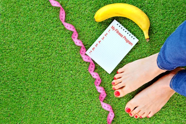 Fitness program and diet concept with a woman legs, measure tape, banana and notebook on a green artificial grass. Copy space.