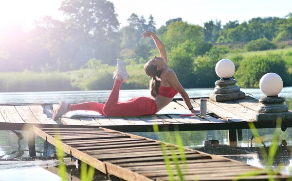 Outdoor training. Morning work-out on a fresh air. Pretty young girl makes yoga exercises on a pier of a pond.